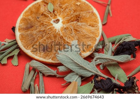 Macro shot of a dried orange slice with sage leaves and hibiscus petals on a red background. The natural mix reflects herbal tea ingredients, organic wellness and aromatherapy aesthetics.