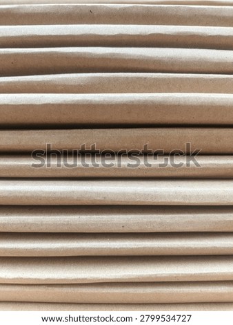 Macro shot featuring a neat stack of corrugated cardboard sheets in creamy brown tones arranged horizontally. Each layer displays a rough yet natural paper fiber texture, with soft shadows between the stacks creating visual depth and a soothing repetitive line pattern. Natural lighting highlights the recycled material surface details and warm earthy nuances. Perfect for eco friendly packaging concepts, paper industry themes, minimalist abstract backgrounds, or sustainability illustrations.