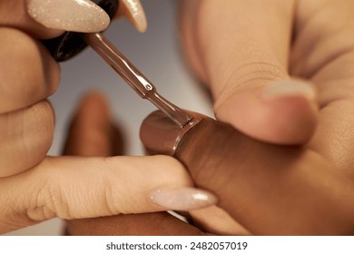 Macro shot of nail tech impeccable work using flat brush covering nail with dark brown lacquer. African American woman getting new nude nails in salon, copy space