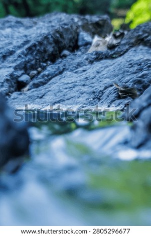 Macro shot of a rough dark grey mountain rock texture with a blurry water background. Showing strong geological lines and details.