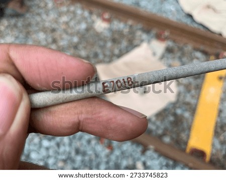 A macro shot of a single E7018 low-hydrogen carbon steel welding electrode. The image clearly shows the "E7018" specification code printed on the flux coating. This type of electrode is widely used in heavy-duty structural welding, pressure vessels, and bridge construction for high-quality, crack-resistant welds. The background features a railway track environment, suggesting industrial maintenance or infrastructure work
