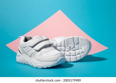 macro, side view one white girl sneaker stands in front of a second sneaker lying on its side with the sole facing the camera on a geometric square paper pink and blue background.