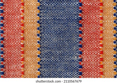 Macro texture of plain weave fabric. Striped cloth background of red, blue, orange and white colors. Textile canvas surface close-up. Design element of colorful woven fabric. Weft and warp. Top view.