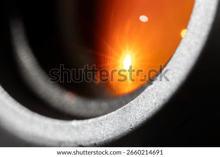 Macro view of a camera lens with a bright sunburst reflection. The star-like flare and bokeh are visible on the orange coated glass, surrounded by the textured metal lens barrel.