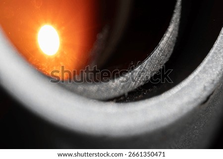 Macro view of a camera lens with a bright sunburst reflection. The star-like flare and bokeh are visible on the orange coated glass, surrounded by the textured metal lens barrel.