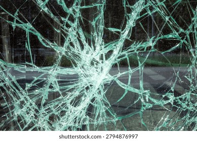 A macro view capturing the intricate, radiating network of cracks spreading from a central impact point across a large pane of shattered safety glass. A detailed close-up photograph showcasing the complex web of fractures across a large sheet of laminated safety glass. The central focus is a dense, pulverised impact zone from which thousands of fine cracks emanate outwards in an intricate, spiderweb-like pattern. These lines intersect to form countless crystalline shards, creating a complex mosaic of broken glass. The transparency and varying angles of the fractured surfaces catch and refract light, producing dazzling highlights and deep shadows that highlight the textures of the damaged material.
Through the clear yet broken glass, the soft-focus (bokeh) background reveals an outdoor environment, with elements like paved ground, green foliage, and perhaps building details visible but heavily blurred and distorted by the fractures, providing context to the scene without distracting from the main subject. The natural light filtering through the scene illuminates the fractures, emphasizing the complex geometric patterns of the shattered panel. This texture and abstract pattern provide a strong sense of texture and scale, suitable for backgrounds, conceptual pieces, or editorial use regarding accidents, security, or fragility. The photograph is captured with high clarity, focusing on the foreground detail while rendering the distant environment as a blurred backdrop.