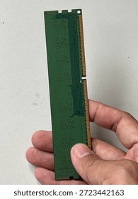 Macro view of green RAM memory module held by hand against a plain background. Detailed shot of desktop computer hardware showing circuit traces and gold contact pins, representing technical repair, hardware upgrade, or system maintenance.