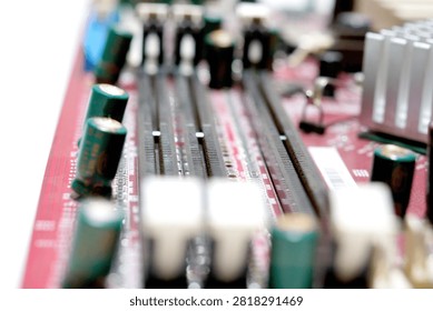 A macro view of a red printed circuit board featuring black DIMM memory slots with white retention clips, green capacitors, and a silver heatsink in a blurred background.