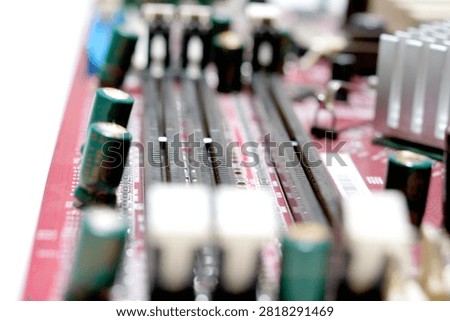 A macro view of a red printed circuit board featuring black DIMM memory slots with white retention clips, green capacitors, and a silver heatsink in a blurred background.