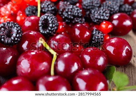Macro view of ripe dark cherries mixed with black raspberries and red currants on rustic wooden board, showing glossy wet skin, vibrant color contrast, shallow depth of field, and fresh natural