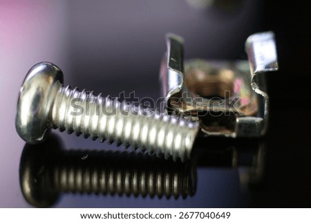 Macro view of a screw and a cage nut - A shiny metal screw lies next to a colorful cage nut, both reflected on a dark, glossy surface.