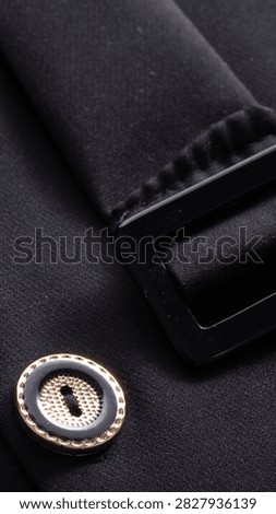 Macro view of textile background showing fine weave of black fabric. Prominent round button displays intricate golden textured center and dark outer ring. Smooth black plastic buckle complements formal aesthetic.