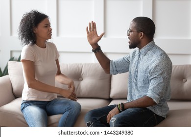 Mad african American young couple sit on couch arguing having family conflict, aggressive furious black husband raise hand intended to hit hurt millennial biracial woman suffer from domestic violence