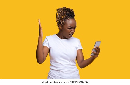 Mad Black Woman Standing On Yellow Studio Background And Looking At Broken Smartphone In Her Hand, Gesturing, Expressing Anger, Panorama With Empty Space. Unhappy African Lady Holding Mobile Phone