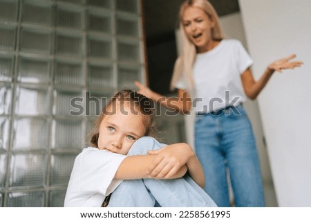 Mad blonde abusive mother emotionally gesturing and shouting at crying little child daughter kid sitting hugging knee, depressed looking away. Concept of scandal, crisis in family, domestic violence.