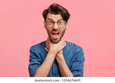 Mad Caucasian male mimicks suicide gesture, keeps hands on neck, frowns face with annoyed expression, wears fashionable shirt, isolated over pink wall. Desperate man suffers from asthmatic attack