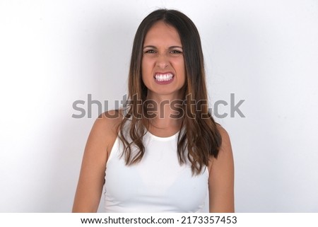 Mad crazy young beautiful caucasian woman wearing white top over white background clenches teeth angrily, being annoyed with coming noise. Negative feeling concept.