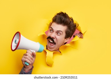 Mad funny male with mustache screaming in loudspeaker and announcing news while looking through torn yellow paper background in studio