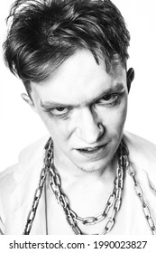 A mad lunatic with makeup and a chain wrapped around his neck screams against a white background. Black and white contrast portrait.
