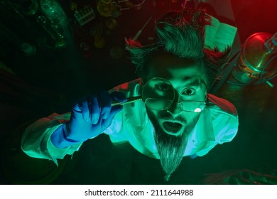 Mad Scientist Looks Through A Magnifying Glass And Is Surprised While Standing In His Laboratory In Green And Red Light. 