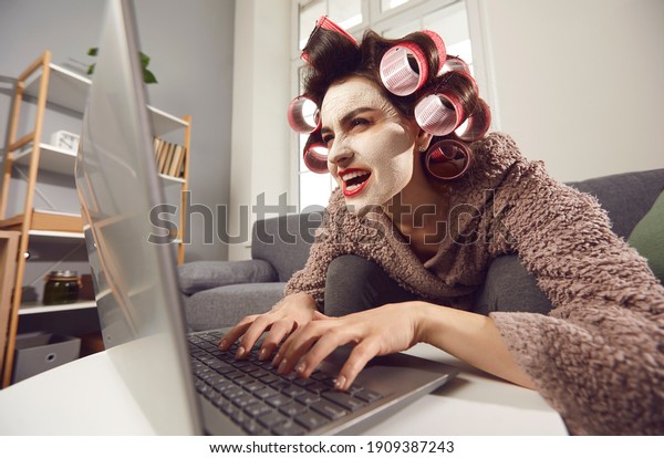 Mad woman in face mask and hair rollers sitting\
looking at laptop computer screen, writing insulting hater comment,\
spreading fake rumour. Jealous wife reading husband\'s direct\
message on social media