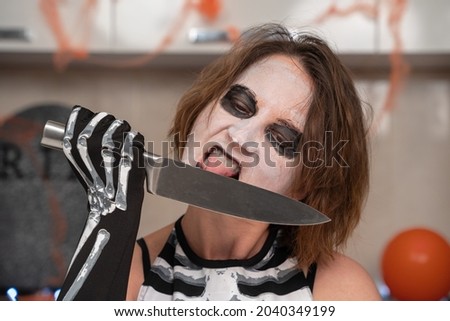 Mad woman in sinister skeleton costume and with creepy makeup in room decorated for Halloween party, front view. She licks sharp knife with crazy look.