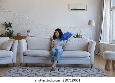 Mad Young Woman Sit On Sofa In Living Room Holding Handheld Blue Paper Fan And Remote Controller From Broken Air Conditioning System Feels Annoyed Suffers From Damaged Device At Hot Summer Day At Home
