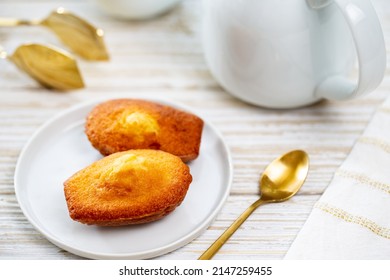 Madeleine cookie or French sponge cake in a white plate alongside a white teapot, golden spoon and tong by a white and gold napkin on white drifted wood background.