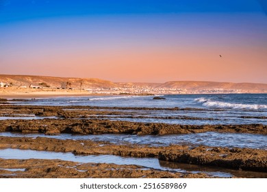 Madraba beach with rugged coastline and crashing waves at sunset. Taghazout town in desert near Agadir, Morocco in background. Rocky coast of Atlantic ocean. Famous surf spot. - Powered by Shutterstock