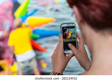 MADRID - MAY 9: Middle-age Woman Taking A Picture With Her Smartphone Of Famous Street Artist Okuda San Miguel As He Finishes A Colorful Ephemeral Graffiti In Madrid, On May 9, 2015.