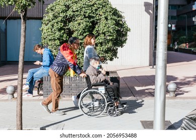 MADRID, SPAIN - OCTOBER 12, 2021:Older Man With The Spanish Flag Around His Neck As If It Were A Cape Walking An Elderly Woman In A Wheelchair During The Military Parade On Spain's National Day