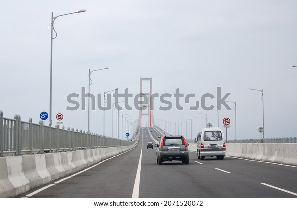 Madura, Indonesia - December\
2011: Scene of the famous Suramadu Bridge and its red suspension\
steel cables with cars and lamp post on road and cloudy sky\
background. 