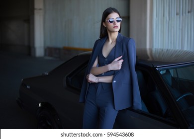 Mafia Lady Outside Japonese Car In The Sea Port. Fashion Girl Standing Next To A Retro Sport Car On The Sun. Stylish Woman In A Blue Suit And Sunglasses Waiting Near Classic Car. 