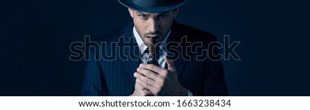 Mafioso lighting cigar and looking at camera on dark, panoramic shot