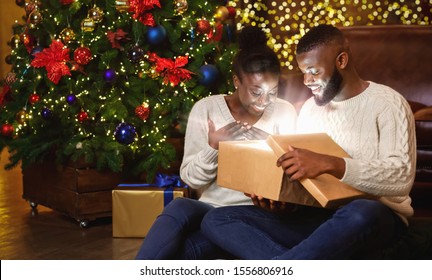 The Magic Of Christmas. Amazed Black Couple Opening Xmas Gift Box With Light Inside, Sitting On Floor Near Christmas Tree At Home