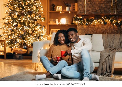 Magic Family Holidays. Cheerful Young Black Couple Celebrating Christmas At Home, Sitting On Floor With Cups Of Coffee, Spending Time Together On Xmas Near Decorated Fir Tree, Copy Space