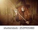 Magic tales. Halloween. Funny forest witch sorceress with red hair, dressed in a black dress and a big hat, stands with her broom in the forest. Fantasy.