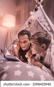 Magical Atmosphere. Happy Positive Nice Man Holding A Flash Light And Smiling While Reading A Book Together With His Son