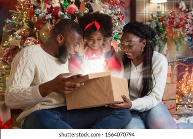 Magical Christmas. Excited African American Family Of Three Opening Shining Gift Box In Living Room Near Xmas Tree, Celebrating Holidays At Home