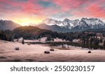 Magical sunrise scenery with mountains, illuminated clouds, a scenic lake and woods on hills in the Alps, frozen meadow and cabins. Beautiful nature in Germany.