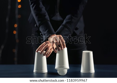 Magician showing tricks with cups on dark background