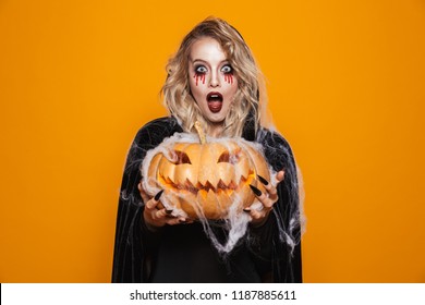 Magician woman wearing black costume and halloween makeup holding carved pumpkin isolated over yellow background