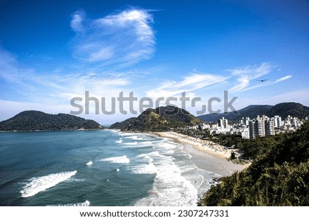 Magnificent aerial view of Praia do Tombo Guaruja Brazil, green mountains, emerald water, strong waves for surfing and birds flying under the blue sky of a beautiful sunny day.