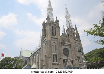 The magnificent Jakarta Cathedral, a grand stone edifice with intricate spires and Gothic architecture, stands prominently under a clear blue sky, framed by green trees on the right. - Powered by Shutterstock - Get 15% off with code: PIKWIZARD15