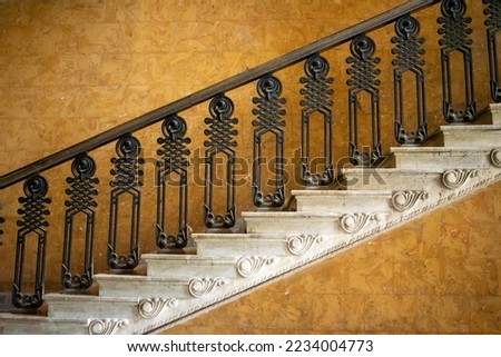 Magnificent marble staircase photographed from the side leads diagonally upwards in front of a yellow wall. Decorated metal railings with art deco style ornaments