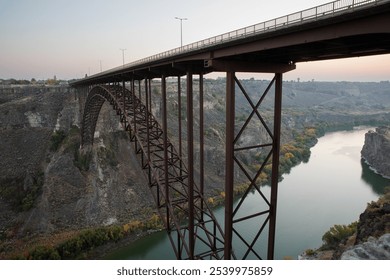 A magnificent steel bridge elegantly stretches across a tranquil river valley at dusk, framed by rugged cliffs and calm waters as seen from twin falls idaho - Powered by Shutterstock