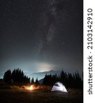 Magnificent view of night starry sky and Milky way over grassy hill with illuminated camp tent and campfire. Concept of hiking, night camping and astronomy.