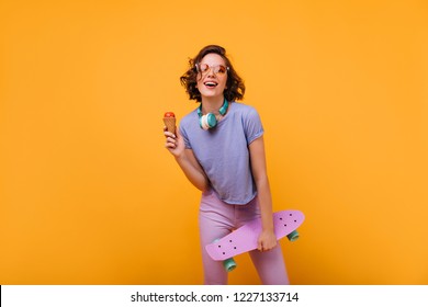 Magnificent white girl in casual clothes posing in headphones. Studio portrait of beautiful lady with skateboard eating ice cream on orange background. - Powered by Shutterstock