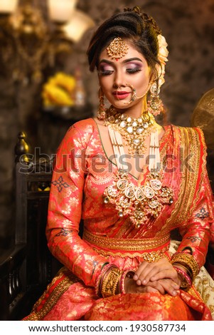Magnificent young Indian bride in luxurious bridal costume with makeup and heavy jewellery sitting in a chair with classic vintage interior in studio lighting. Wedding fashion.