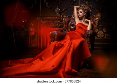 Magnificent young woman in luxurious red dress and precious jewelery is sitting in a chair in a luxury apartment. Classic vintage interior. Beauty, fashion.